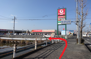 澄み渡る青空の下、大きな駐車場を備えたショッピングセンターの風景を捉えており、手前の歩道から左側にある橋を渡って店舗の手前の細い通路へと進む経路を、太い赤い矢印で示している写真