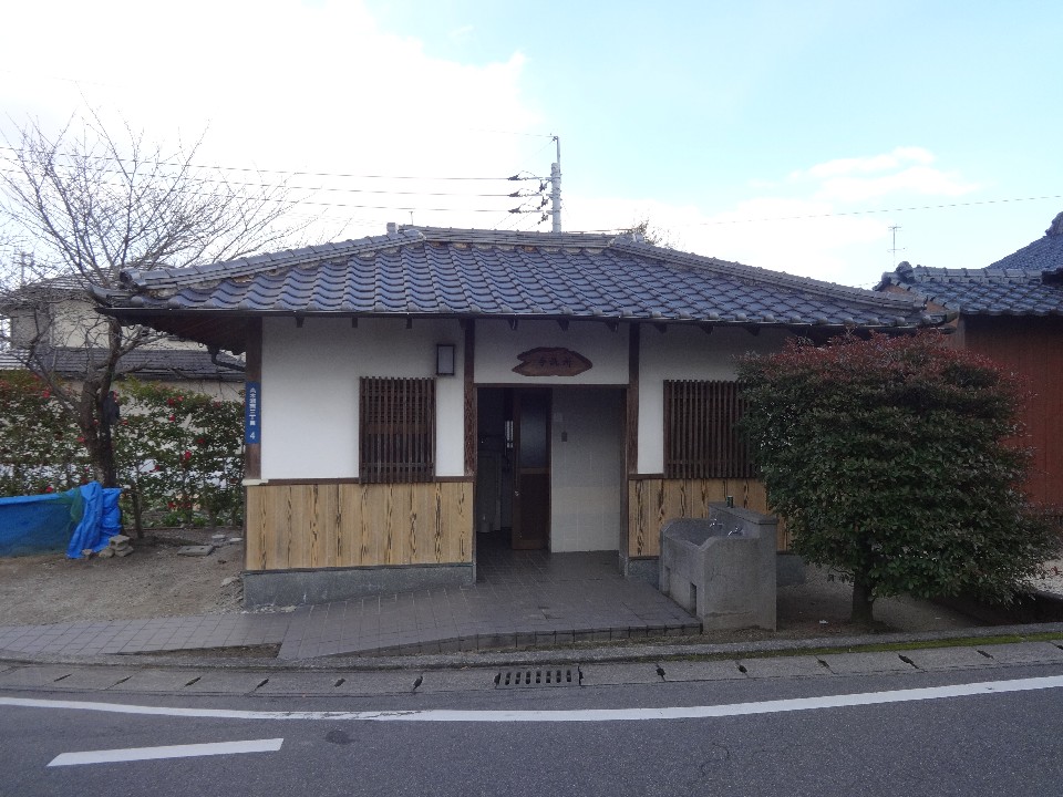 瓦屋根と白い壁、木目調の腰壁が特徴的な和風建築の公衆トイレが道路沿いに建っており、入口横には石造りの手洗い場や整えられた植栽、格子窓が配置された、落ち着いた色合いの平屋のトイレを正面から捉えた写真