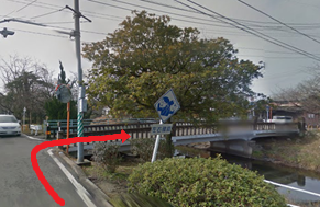 青い標識や低い植栽が配置された、電柱や大きな木が立つ細い道路から、川に架かるコンクリート造りの「植木橋」を右折して渡る経路が赤い矢印で示されている写真