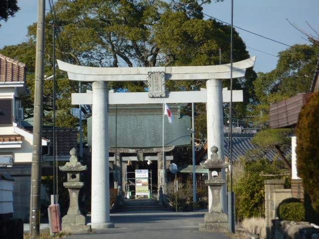 大きな楠の緑を背に、堂々とした石造りの明神鳥居が参道をまたぐように立ち、その奥に社殿の屋根や翻る日の丸、そして左右に並ぶ石灯籠を正面から捉えた、晴れやかで威厳に満ちた神社の風景を捉えた写真