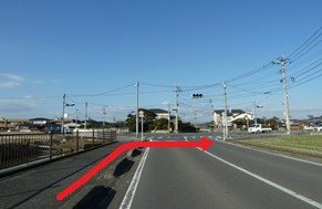 青空の下、周囲の住宅街や遠くの山々を背景に、電柱が並ぶ二車線の道路において、手前の歩道側から車道の中央へと進み、さらに前方の交差点で右折することを示す赤い矢印が示されている写真