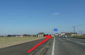 広大な空と田園風景が広がるのどかな住宅街の景色を背景に、見通しの良い直線道路の歩道から、前方の青い道路標識がある付近で左側の脇道へと曲がるルートを示す赤い矢印が重ねて描かれている写真