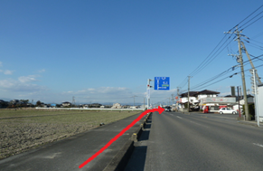 晴天の下、民家が並ぶのどかな郊外の風景を背景に、左側に広がる田畑に沿った歩道から車道へと斜めに進み、前方の青い案内標識がある付近で右折するルートを赤い矢印で示した写真