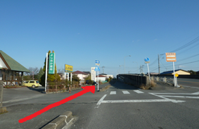 左側に薬局や看板が並ぶ建物があり、正面に横断歩道やスロープ状の道が見える交差点において、手前から直進して細い脇道へと進むルートを赤い矢印で描いた、澄み渡る青空の下の穏やかな街並みを写した写真