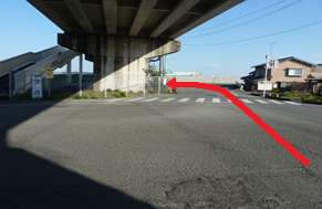 快晴の空の下にある静かな住宅街の風景を背景に、高架下の影が落ちる交差点において、手前から横断歩道の方へと進み、コンクリート製の巨大な橋脚の脇を抜けて左奥の細い道へと続くルートを赤い矢印で示した写真