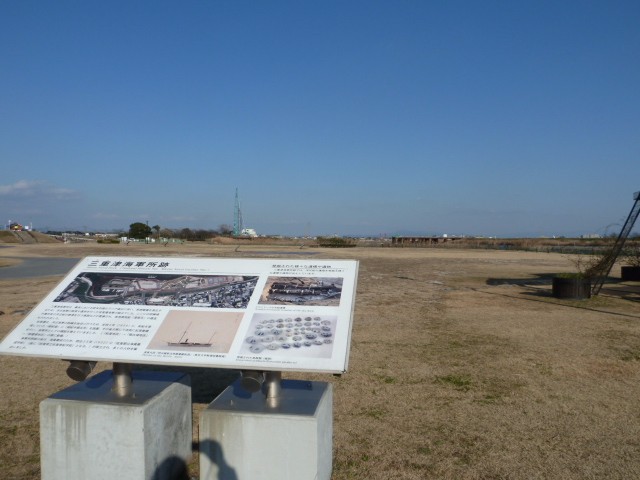 抜けるような青空の下、広大な芝生広場の手前に設置された「三重津海軍所跡」の解説パネルを主役に、遠くに見えるクレーンや平坦な景色を背景として、歴史遺構の静かな佇まいを写した写真