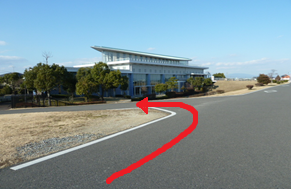 抜けるような青空の下、周囲に芝生や木々が広がる開放的な施設が見える背景に、舗装された広い道路のカーブに沿って左奥の近代的な建物の方へと進むルートを赤い太い矢印で示した写真