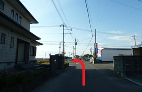 強い日差しが降り注ぐ中、静かな郊外の街並みの住宅の間を抜ける細い道を進み、前方の交差点を左に曲がって白いフェンスが続く道へと入るルートを赤い矢印で示した写真