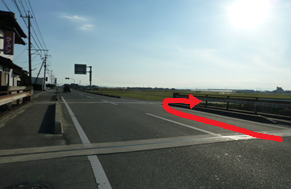抜けるような青空の下、のどかな郊外の街並みと遠くの山々を背景に、民家が並ぶ二車線の道路において、手前から真っすぐ前へ進み、その先の交差点を右折するルートを赤い矢印で示した写真