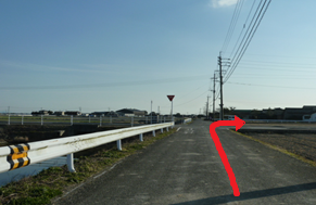 強い日差しの中、田園風景が広がるのどかな景観を背景に、左側に続く水路と白いガードレールのある直線道路において、前方の止まれの標識がある交差点を右折して民家が並ぶエリアへと進むルートを赤い矢印で示した写真