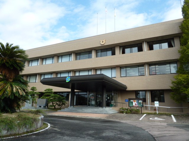 青い空の下に建つ3階建ての重厚な茶色の外壁の建物で、正面玄関の黒いひさしには緑と青のロゴマークが掲げられており、周囲には南国風の樹木や松の木が植えられ、手前にはアスファルトの通路が広がる佐賀市役所 東与賀支所の外観写真
