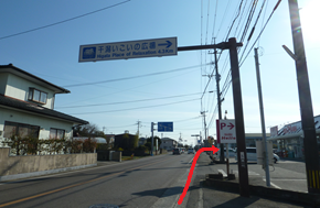 晴れ渡った空の下、住宅が並ぶ道路上に「干潟いこいの広場」への案内標識が立ち、右手にある店舗の駐車場入り口を示す看板へ向かって赤い矢印が書き込まれ、遠くまで続く道と電柱が並ぶ風景を捉えた写真