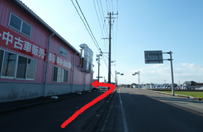 左手に「中古車販売」と書かれたピンク色の大きな倉庫風の建物が建つ直線の道路で、建物の脇を通って奥へと進むルートを示す太い赤色の矢印が路面上に示された写真