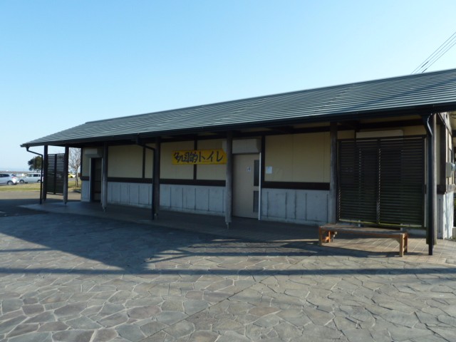 青空の下、石畳の広場に面して建つ平屋建ての和風建築で、壁面には黄色い看板に黒文字で「多目的トイレ」と大きく書かれており、建物の前には木製のベンチが置かれ、落ち着いた外観を持つ公共トイレの写真