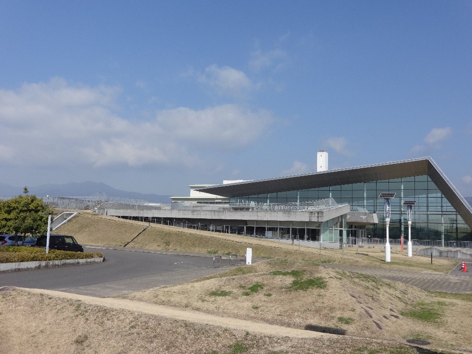 手前には舗装された道路や駐車場が広がり、遠くには霞んだ山々が連なるなだらかな芝生の丘に沿って建てられた、大きなガラス張りの壁面と鋭角な屋根が特徴的なモダンなデザインの広大な佐賀市健康運動センターの外観を捉えている写真