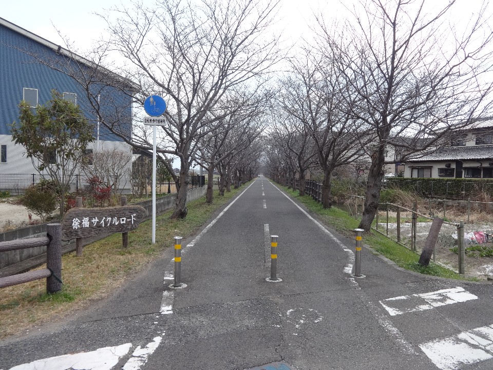 葉の落ちた桜並木がどこまでも続く真っ直ぐな舗装路が伸びており、入り口には自転車歩行者専用の標識と進入禁止のポールが並ぶ横に、「徐福サイクルロード」と刻まれた木製の看板が立っているサイクリングロードの写真