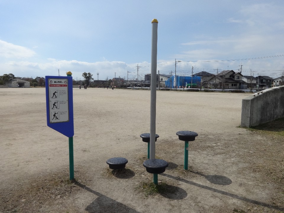 晴れ渡る空の下、広い砂地の広場に設置された「あしのばし」と書かれた青い看板付きの健康遊具と、その周囲に配置された高さの異なる4つのステップや中央のポールを、遠くの住宅街を背景に捉えた写真
