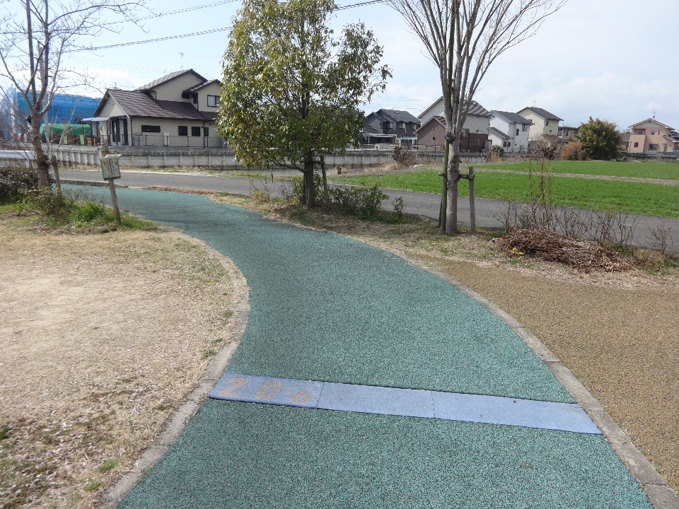 薄曇りの空の下、手前に「200」と刻まれた青いラインがある緑色の舗装路が緩やかに左へカーブし、その先に並ぶ若木や住宅街、そして隣接する青々とした田畑の風景を捉えた写真