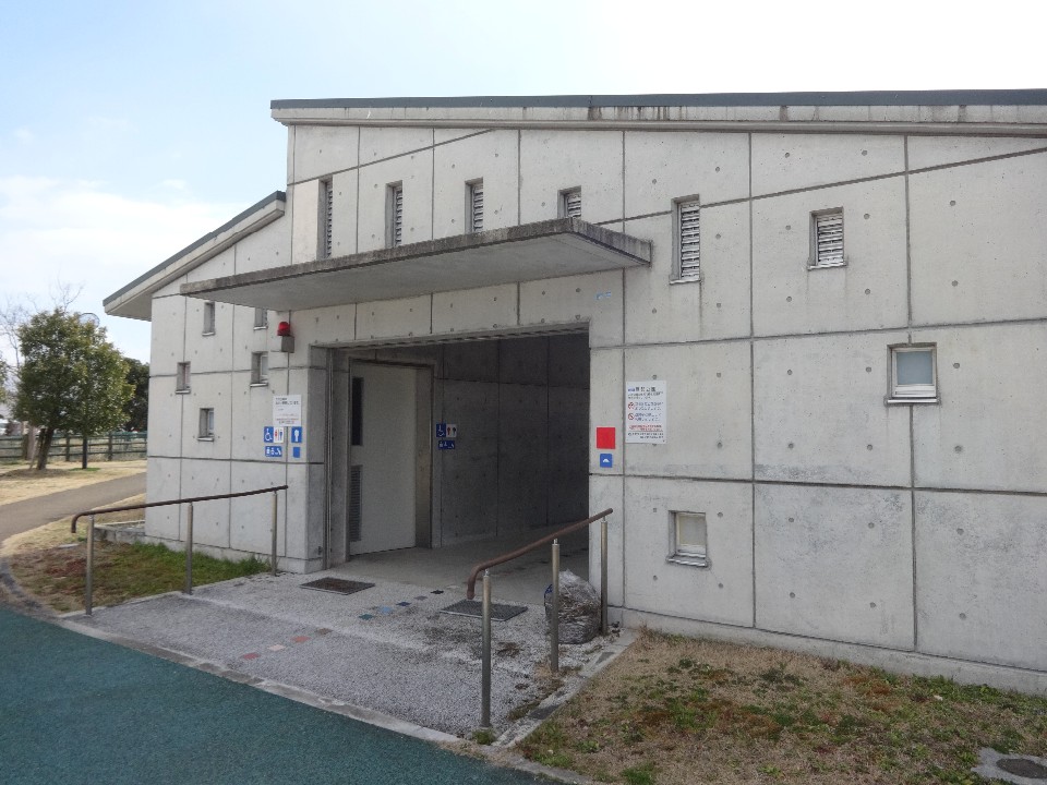 晴れ渡る空の下、コンクリート打ち放しの壁面に小さな窓が並ぶ公衆トイレの建物を捉え、入口には手すり付きのスロープや多機能トイレを示す案内板が設置された、バリアフリーに配慮された施設の外観の写真
