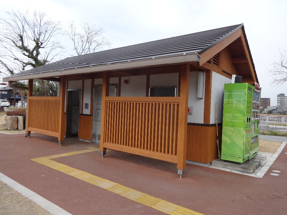 曇り空の下で、温かみのある木材と白い壁面を組み合わせた和風モダンの公衆トイレが建っており、建物の右端には黄緑色の飲料自動販売機が、手前の地面には視覚障害者用の誘導ブロックが敷設されている公園内の施設を捉えた写真