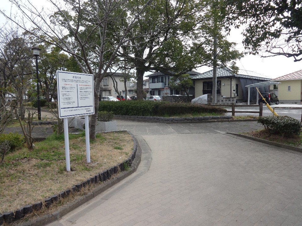 曇り空の下、舗装された遊歩道の脇に公園の利用規則が書かれた白い立て看板が設置されており、その奥には住宅街や青々と茂った木々が並ぶ静かな公園の入り口付近を捉えた写真