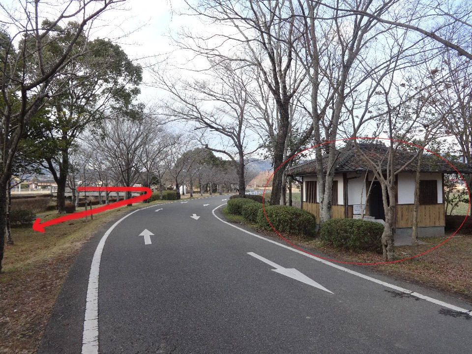 緩やかに右へカーブするアスファルトの道路沿いに、瓦屋根の小さな平屋の建物が赤い円で強調されており、周囲には葉の落ちた木々や低い植栽が並び、左側の草地には進行方向を示す赤い矢印が描かれた、公園内の風景の写真