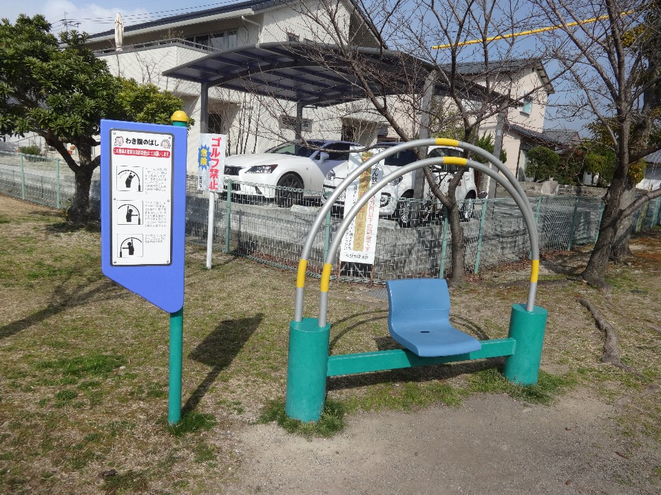 住宅街の公園内に設置された、青い一人掛けの座面と背後に円弧状の金属フレームを備えた健康遊具「わき腹のばし」と、その左側に立てられたイラスト入りの使用説明看板が設備を詳細に示す写真