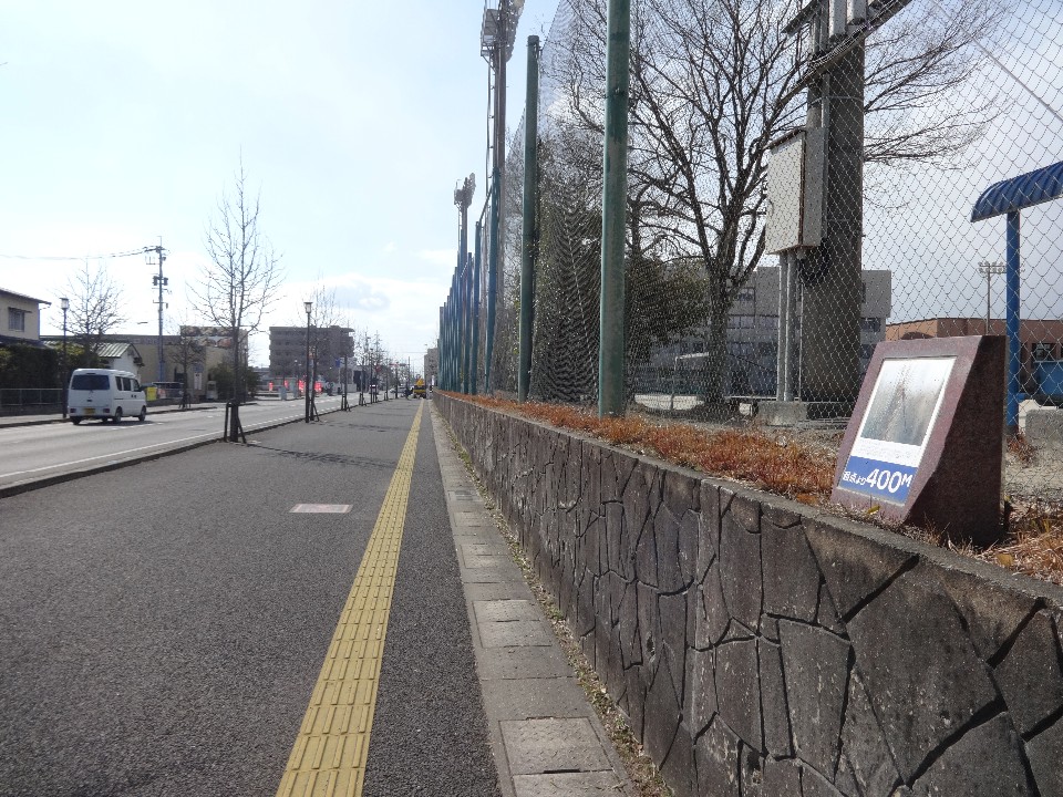 点字ブロックが設置された平坦な歩道の右側に、「アオモンイトトンボ」の解説と「起点より400メートル」の文字が記された石造りの案内プレートが置かれ、その背後には高いネットが張られたスポーツ施設や街路樹が並ぶ、屋外の遊歩道の風景を捉えた写真