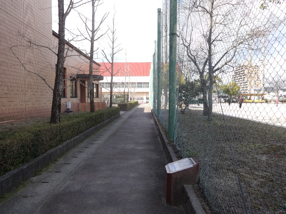 左側にタイル貼りの建物、右側に高いネットに囲まれたグラウンドがある細い舗装路の風景で、手前の路肩には「ハグロトンボ」の解説と「起点より600メートル」と記された石造りの案内プレートが設置されている、公園内の遊歩道の様子を捉えた写真