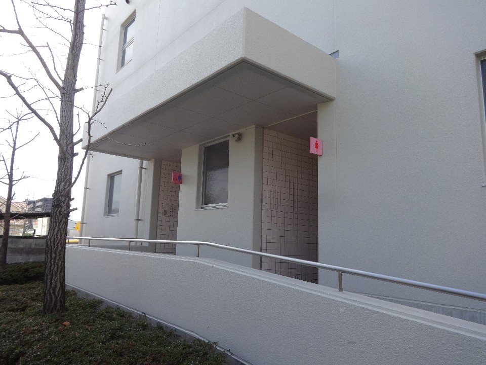 白い壁の建物に設置されたスロープの先に、ピンク色の背景に女性のシルエットが描かれた女子トイレの案内看板が複数掲示されており、バリアフリーに配慮された施設入口の写真
