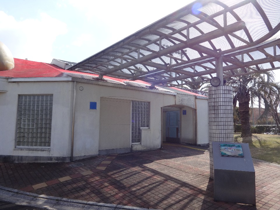 日差しが降り注ぐ中、白い外壁にガラスブロックがはめ込まれた平屋建ての建物の入口付近で、金属フレームの大きな日除け屋根がエントランスを覆うように設置されている写真