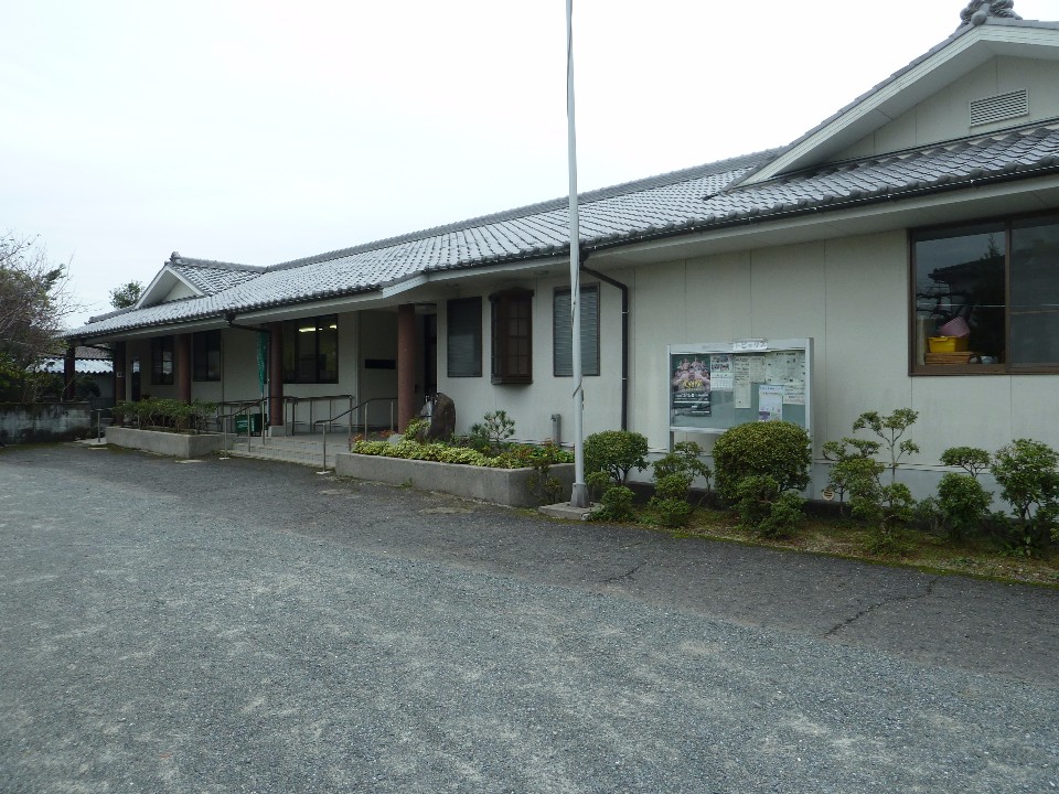 平屋建ての瓦屋根が特徴的な和風建築の建物外観と、その手前に広がる砂利敷きの広場、壁際に設置された掲示板や植栽、奥へと続くスロープ付きの入り口周辺の様子を詳細に捉えた、落ち着いた雰囲気の蓮池公民館の写真