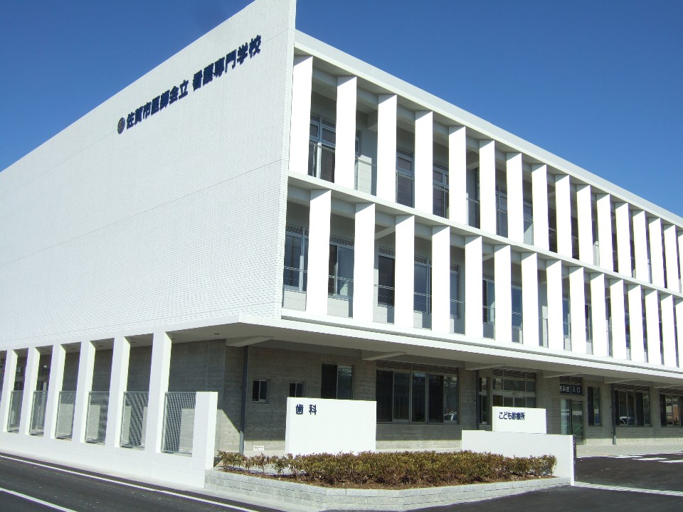 白い外壁と縦長の窓が整然と並ぶ三階建ての近代的な建物で、前面には低木の植栽が整えられた清潔感のある佐賀市休日夜間こども診療所の外観写真