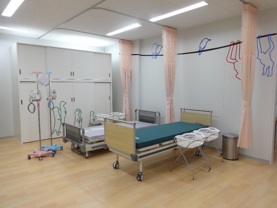 白い壁と木目調の床を持つ明るい室内に、カーテンで仕切られたスペースに2台の病院用ベッドと点滴スタンド、折りたたみ式のランドリーバスケットが配置され、壁には動物のシルエットが描かれたデザインが施された佐賀市休日夜間こども診療所 処置室の写真