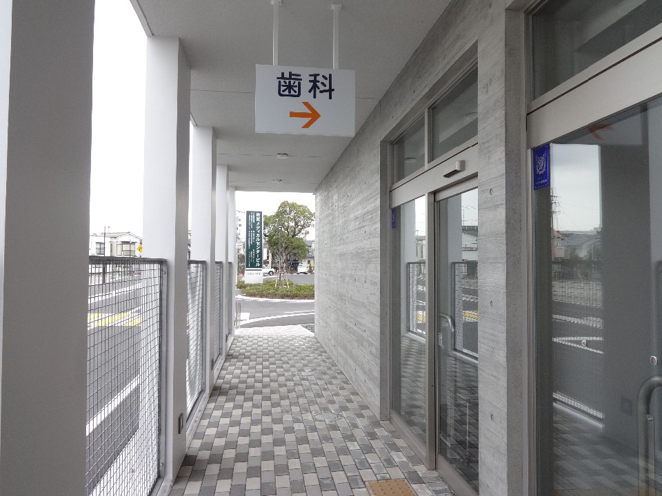 灰色のタイルが敷かれた屋外通路に、右側にはガラス扉とコンクリート壁が並び、天井から「歯科」と書かれた案内板がオレンジ色の矢印付きで吊り下げられている佐賀市休日歯科診療所入口の写真