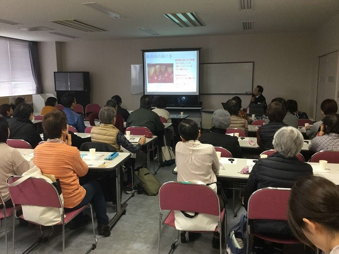 教室内で複数の人が机に座り、前方のスクリーンに歯周病に関するスライドが映し出され、講師が説明をしている様子で、参加者は資料や飲み物を手元に置いて講義を受けている写真
