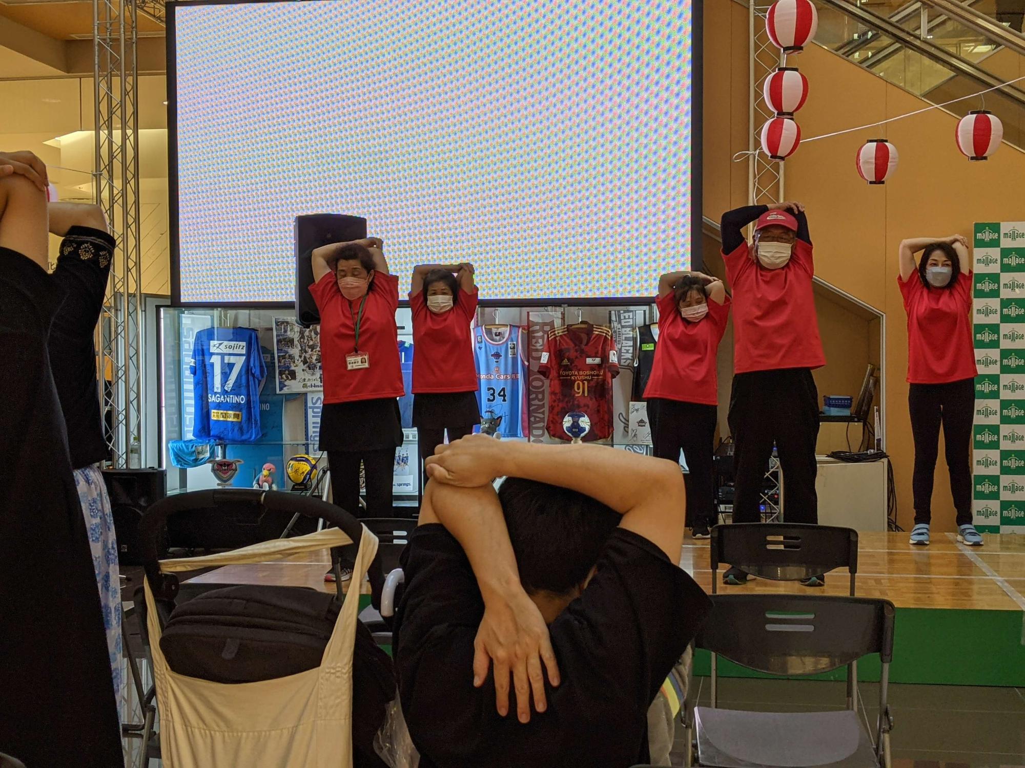 屋内のイベント会場で、ステージ上に赤いシャツと黒いパンツを着た複数の人々が並び、両腕を頭の後ろに回すストレッチを実演しており、前方の観客も同じ動作をしている体操の様子を写した写真
