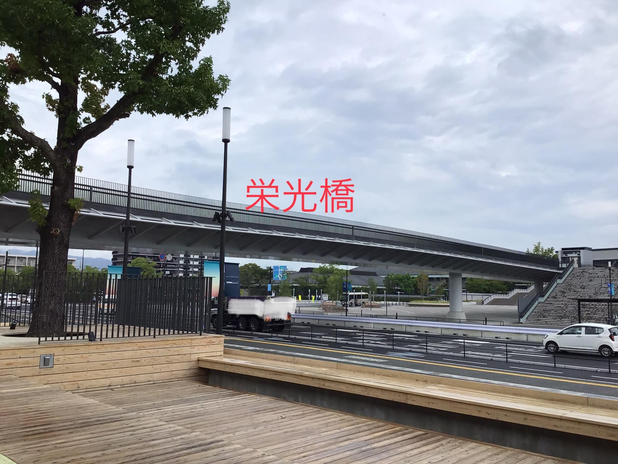 「栄光橋」という赤い文字が添えられた、近代的なデザインの巨大な歩道橋が道路をまたぐように架かり、手前には温かみのある木製デッキの広場が広がる、開放感のある都会的な公共空間を捉えた写真
