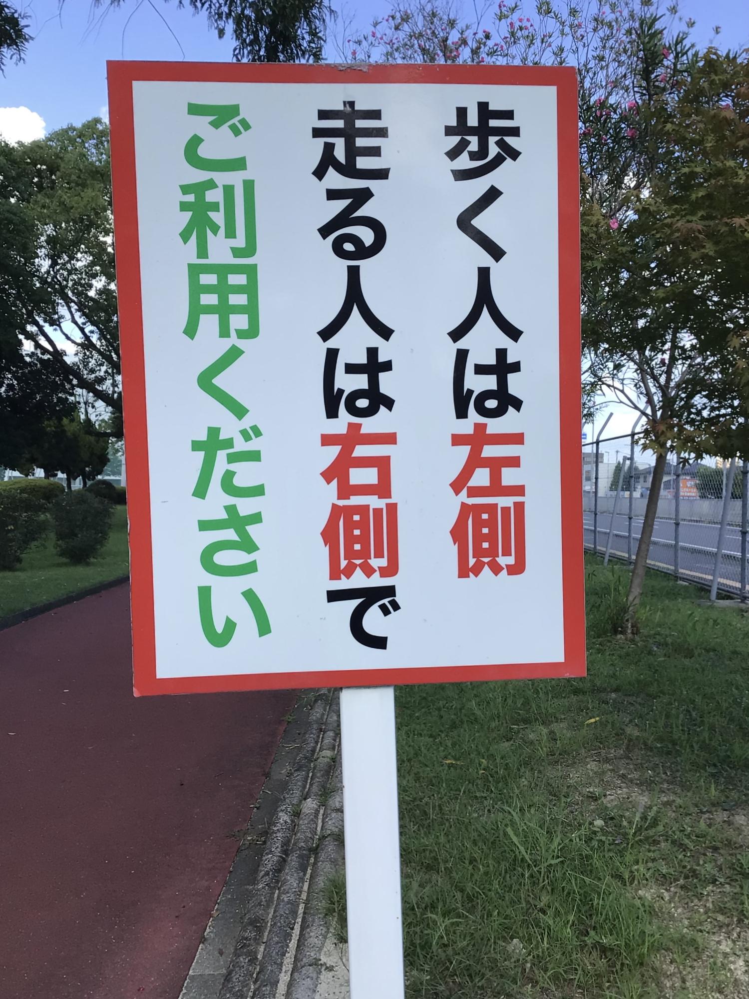 赤枠で縁取られた白い看板に、「歩く人は左側 走る人は右側でご利用ください」と大きく黒と赤の文字で記され、背後には赤茶色のランニングコースが木々の緑に沿って続く、利用ルールを明示したコース脇の標識の写真