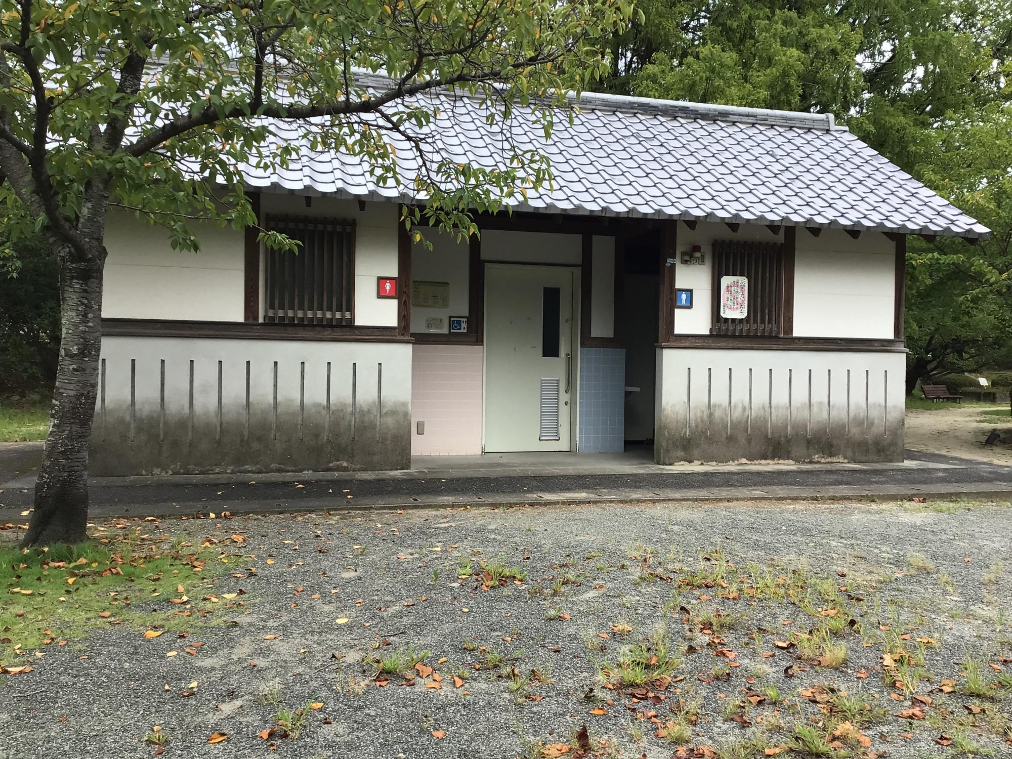 瓦屋根を冠した白壁の平屋建てが、木々の緑に囲まれた静かな広場に佇み、建物の正面には多機能トイレを含む複数の入り口が並ぶ、公園内の和風な外観の公衆トイレの写真