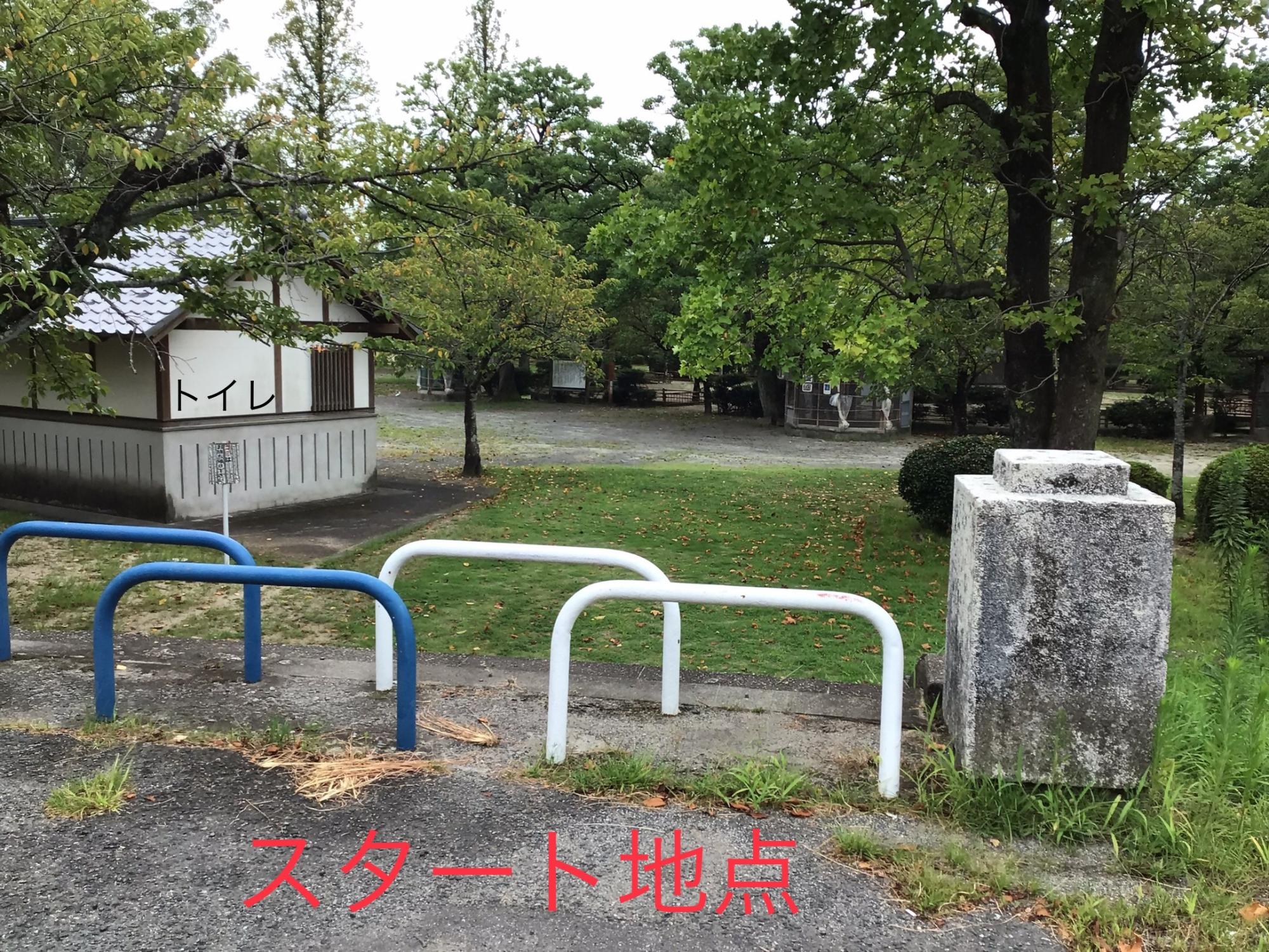 「スタート地点」と赤い文字で記されたアスファルトの道から、白と青の車止めが並ぶ先を見渡し、左手には「トイレ」と書かれた清潔感のある建物、奥には緑豊かな木々と芝生の広場が広がる、公園の入り口付近の風景を捉えた写真