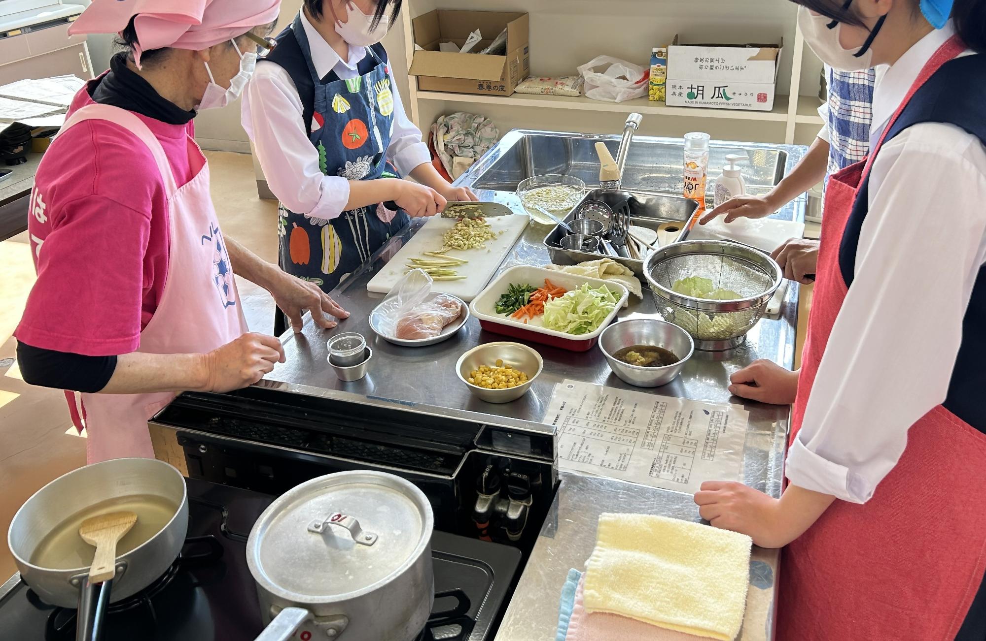 調理室のステンレス製作業台を囲み、エプロンとマスクを着用した女性たちが、まな板で野菜を細かく刻んだりボウルに具材を準備したりしながら、レシピを確認して協力して調理を進めている様子を捉えた写真