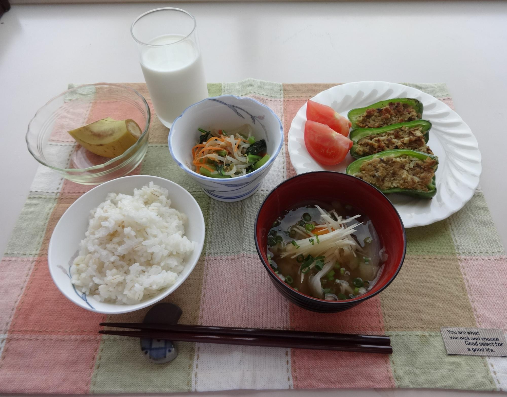 チェック柄のランチョンマットの上に、白米、具だくさんの汁物、ピーマンの肉詰めとトマト、野菜の和え物、カットされたバナナ、コップ一杯の牛乳がバランス良く並べられた、健康的で彩り豊かな和食膳の写真