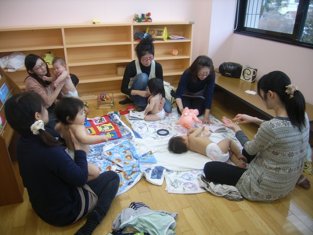 木製の棚が並ぶ室内で、床に敷かれたタオルや布の上に複数の乳幼児が座ったり寝かされており、周囲には大人が円形に座ってベビーマッサージを行っている写真
