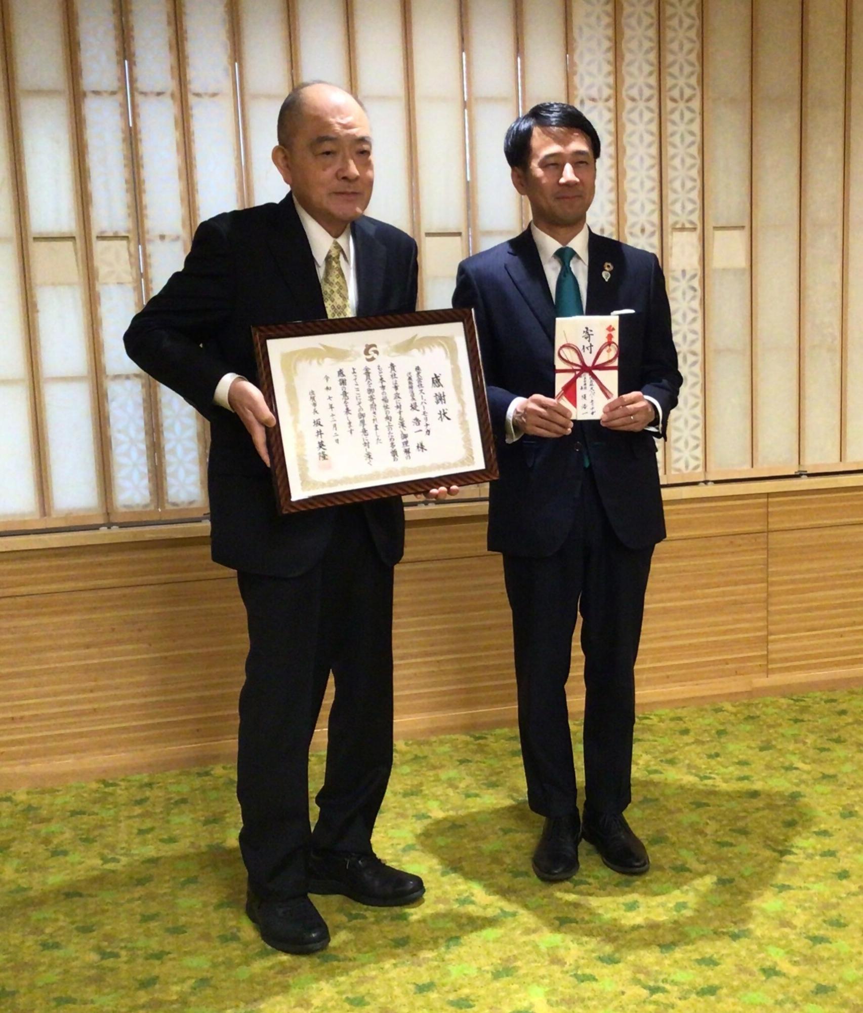 左側の男性が「感謝状」と書かれた額縁を大切そうに掲げ、右側の市長が目録を手に持ちスーツ姿で並んでいる記念撮影の写真
