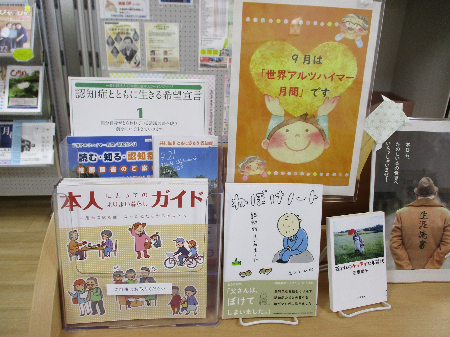 図書館の棚に、9月は「世界アルツハイマー月間」であることを知らせるオレンジ色のポスターや、「本人にとってのよりよい暮らしガイド」などの認知症に関連する複数のパンフレットや書籍が整然と並べられている写真