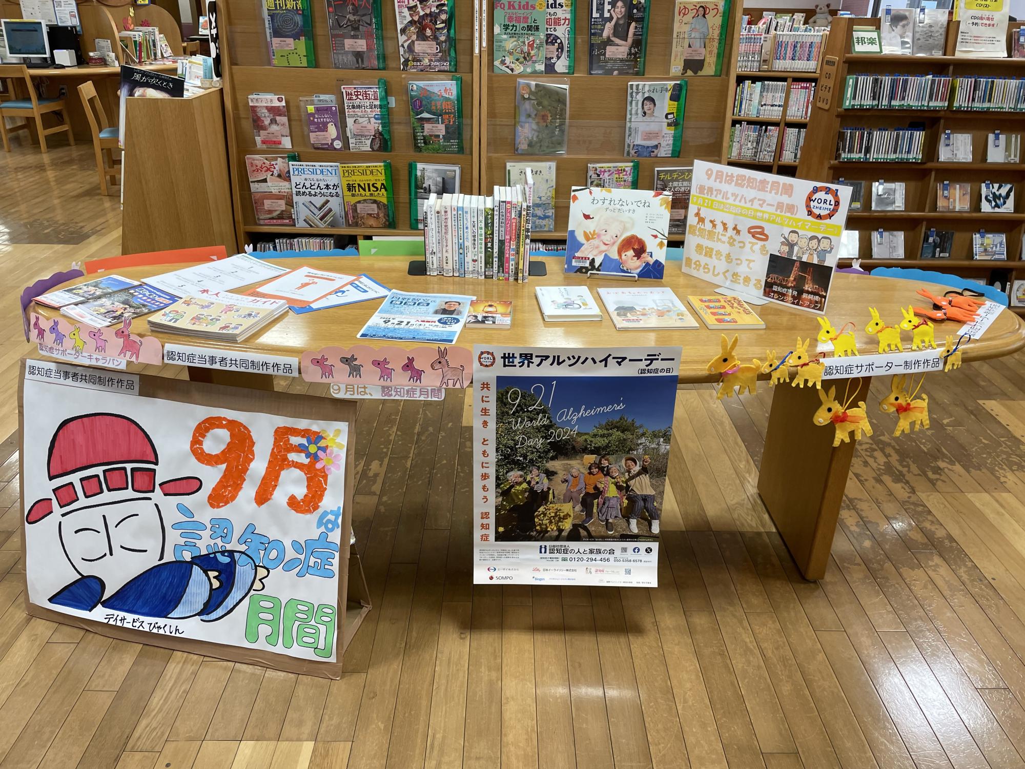 楕円形の木製テーブルに、世界アルツハイマーデーのポスターや認知症関連の書籍が並び、手前には「9月は認知症月間」と書かれた手描きの大きな掲示物やロバの形をした黄色の手作り飾りが添えられた特設コーナーの写真