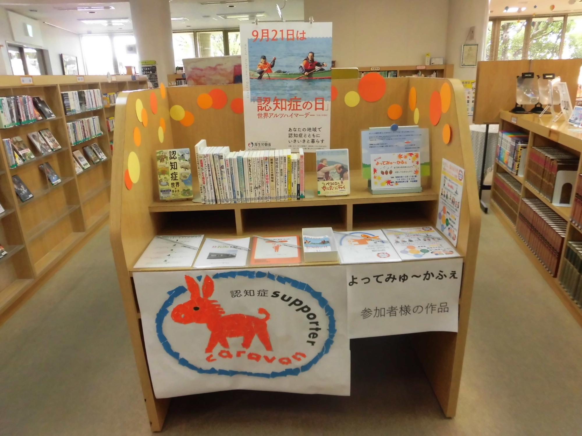 図書館の棚に、世界アルツハイマーデーのポスターや認知症関連の書籍が並び、手前には「佐賀市認知症ケアパス」や介護施設のパンフレット、認知症カフェの案内やカフェ参加者が作成されたチラシが並んでいる。また、認知症サポーターキャラバンのロゴであるオレンジ色のロバのイラストが描かれた横断幕が掲げられている写真。