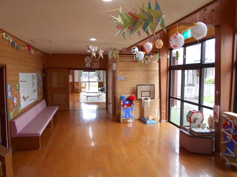 天井から星形の飾りや吊り下げモビールが揺れる明るくて広い廊下に、ガラス窓沿いの遊具や掲示物、奥の棚や机が並ぶ華やかな室内空間の写真