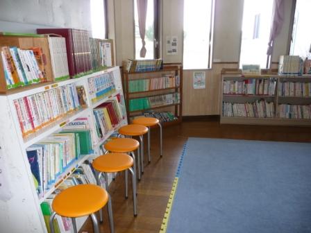 本棚に多くの児童書が並び橙色の丸椅子と広いマットスペースが配置された明るい図書コーナーの写真